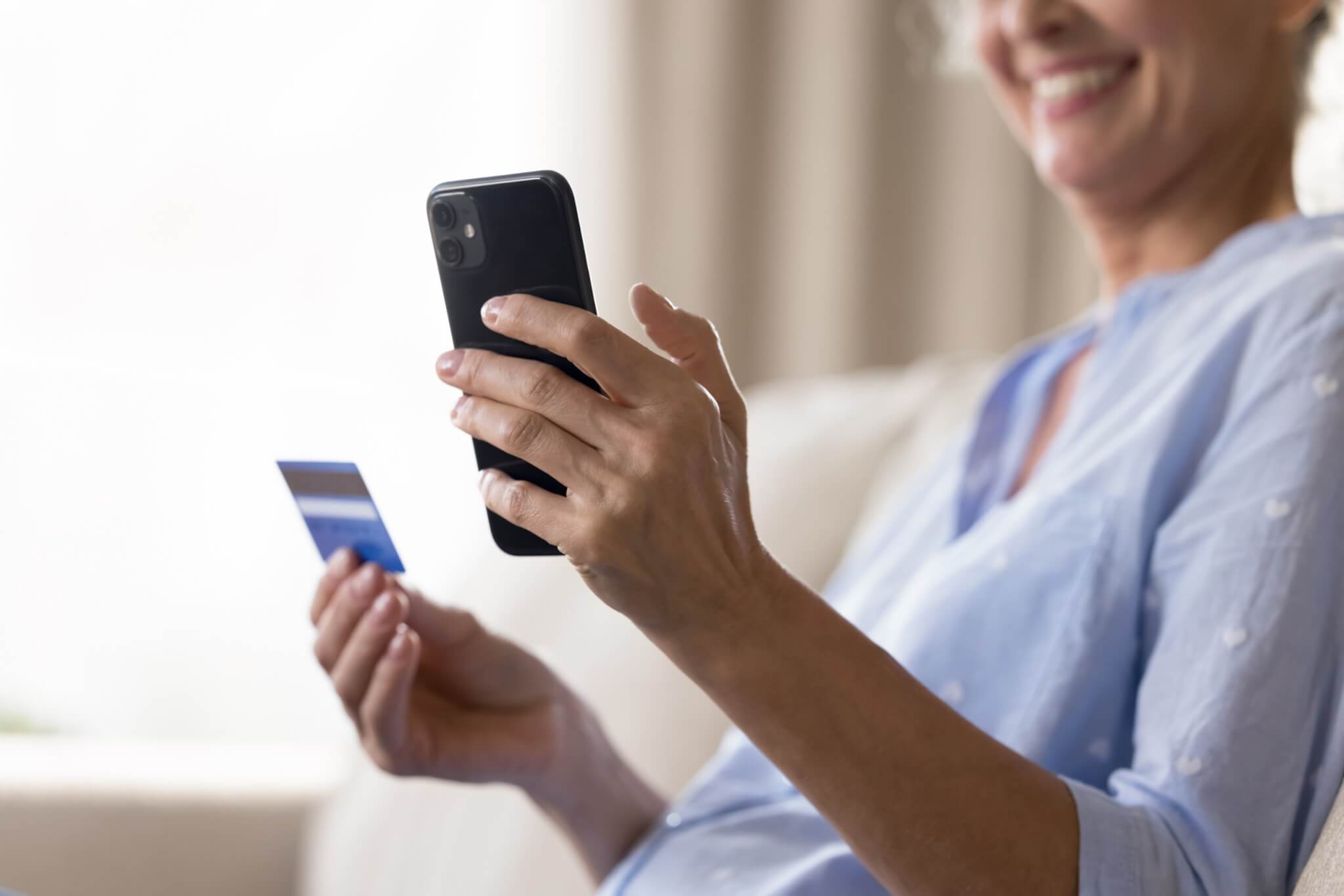 Close up cropped shot smiling aged female holding smartphone credit bank card in hands.
