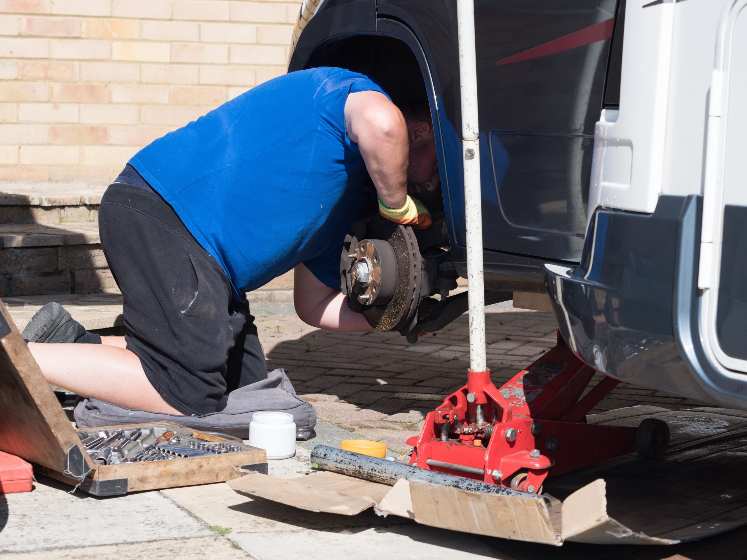 A mechanic has jacked up a motorhome recreational vehicle and removed a wheel and peers under the wheel arch checking the brakes.
