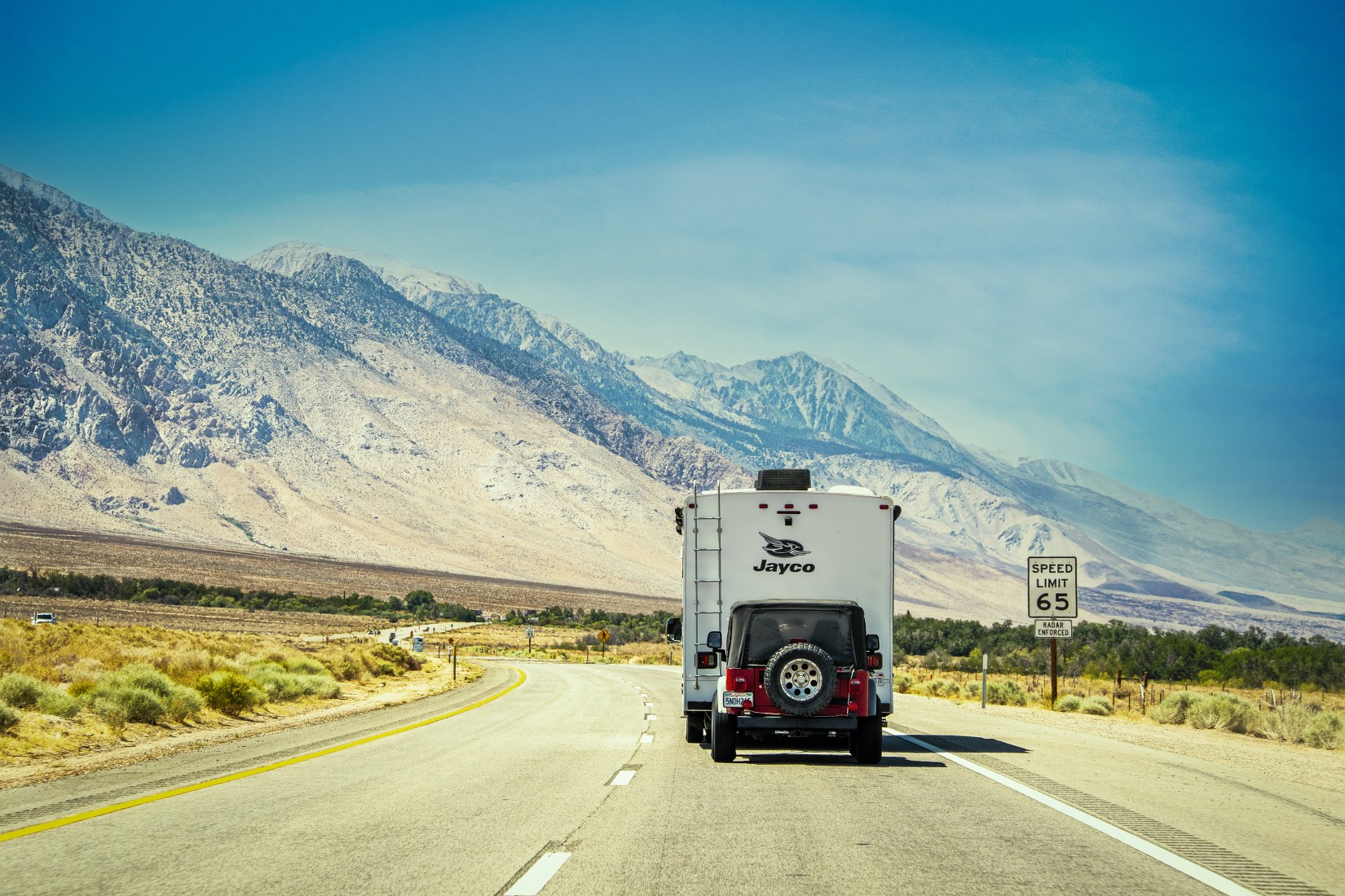 Camper towing Jeep drives on curving highway with Sierra Nevada mountains in background in arid environment of Eastern California.