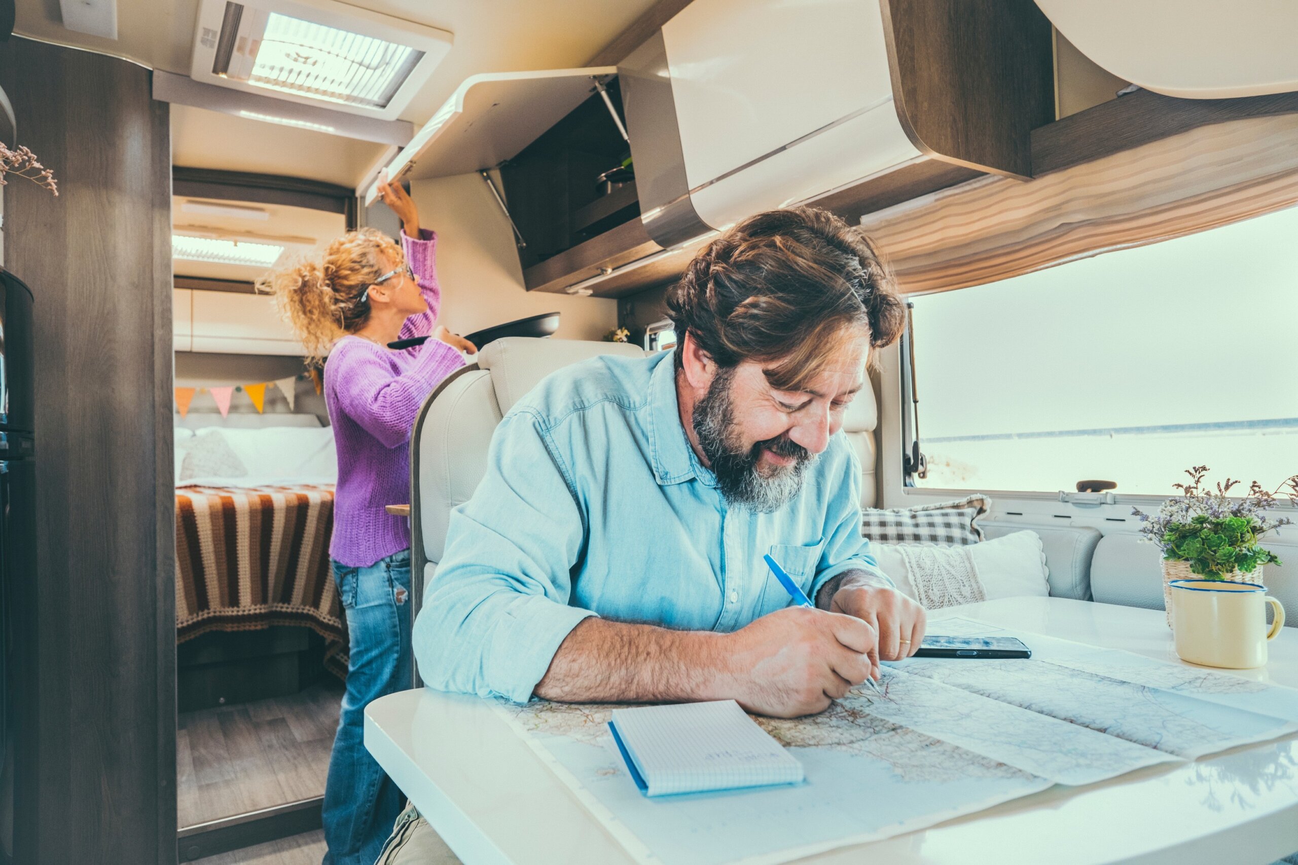 Bearded man planning next destination travel holiday vacation sitting at the table inside his camper van. Woman in background cooking and managing storage.