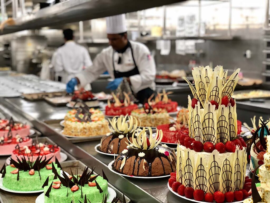 Big selection of cakes on a cruise ship. Big galley with many chefs.