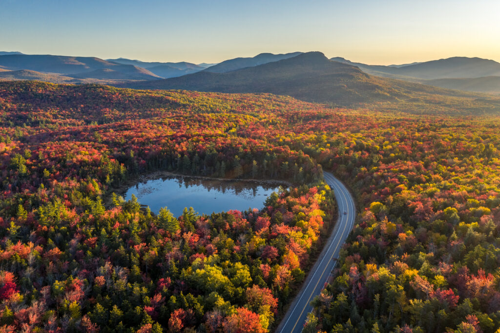 A road, highway through a forest with trees in fall colors of red, orange and yellow, lake, mountains, Kancamagus Highway, White Mountains, New Hampshire.