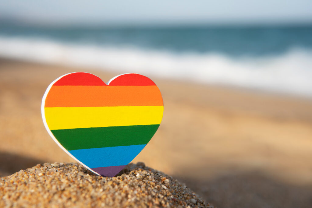 Rainbow Gay Pride heart sign on the sand with the sea on background. LGBT and human rights minimal concept. Gender equality concept. Copy Space. Romantic trip gay honeymoon. Same-sex couple.