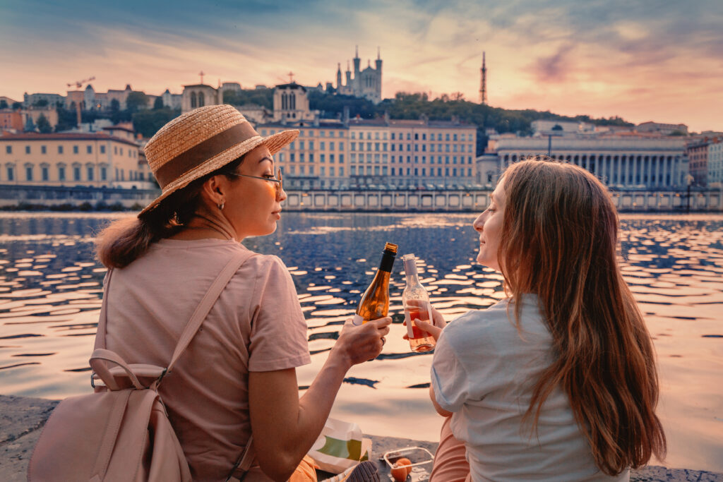 Two happy girls close friends celebrating and toasting and drinking wine from a little bottles and watching a sun goes down over Saone river in Lyon city, France