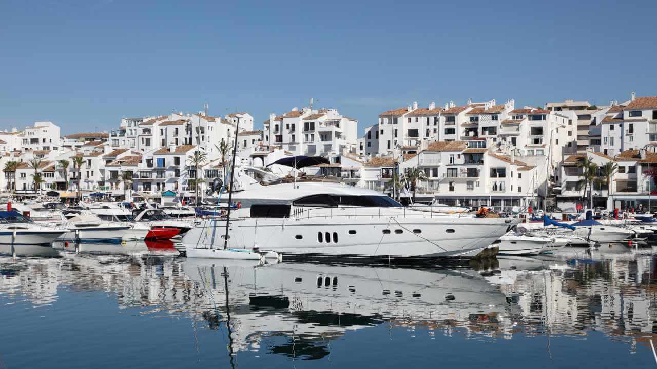 Several boats are docked in a marina with white buildings in the background.
