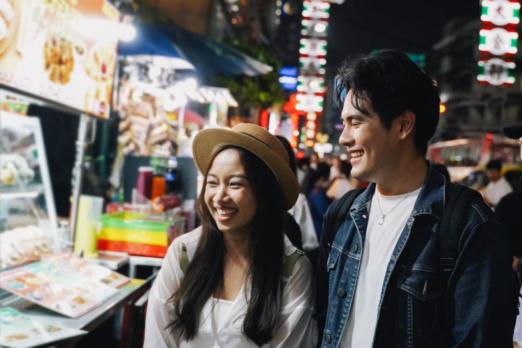 Asian couple tourist backpacker enjoying and eating street food in night market with crowd of people at Yaowarat road, Bangkok