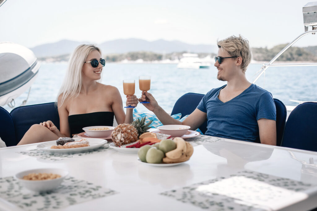 Charming adult couple man and woman are having dinner at the table of their huge and expensive white yacht, smiling and holding glasses of juice in their hands, enjoying each other's company