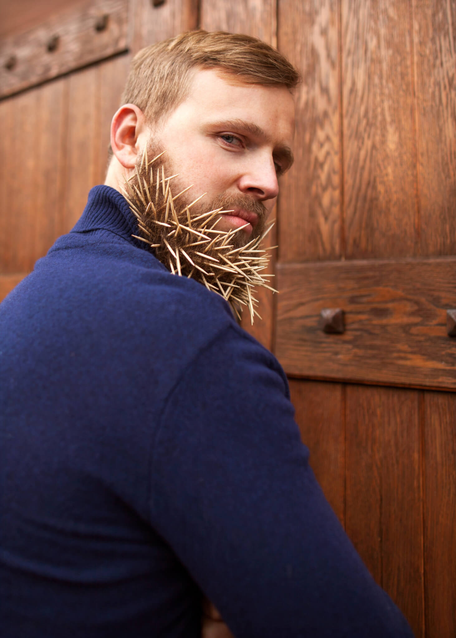 fashion hipster guy standing near a wooden door toothpicks in his beard, hatred, anger, contempt, deceit, greed.