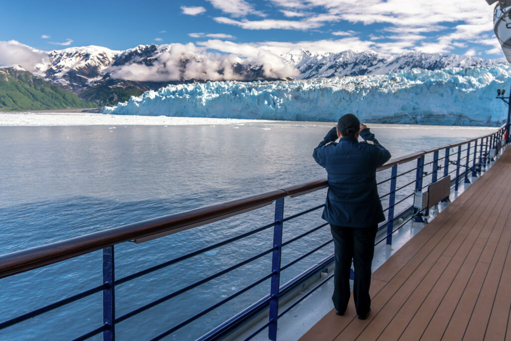 Woman is wearing a stripes and taking a picture of Hubbard Glacier on her mobile phone. Lady is crew member on a cruise ship. Working as a ships officer. Snowy mountains and iceberg.