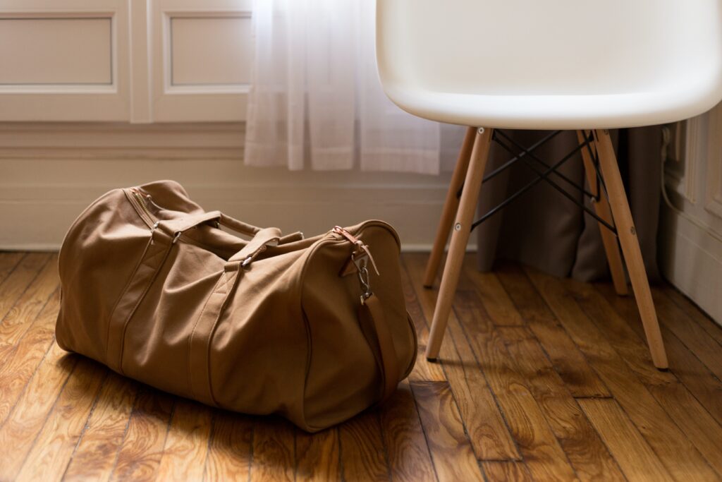 A leather and canvas sand-colored duffel bag. An Eames chair in white is in the background.