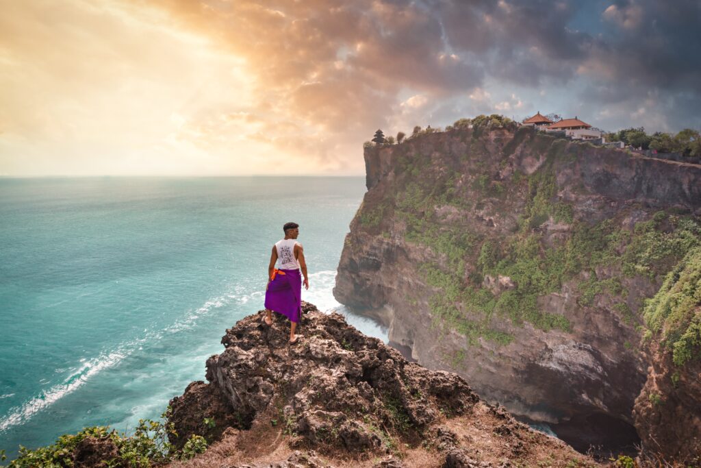 Guy standing on cliff at uluwatu temple in Bali Indonesia during a beautiful golden sunset.