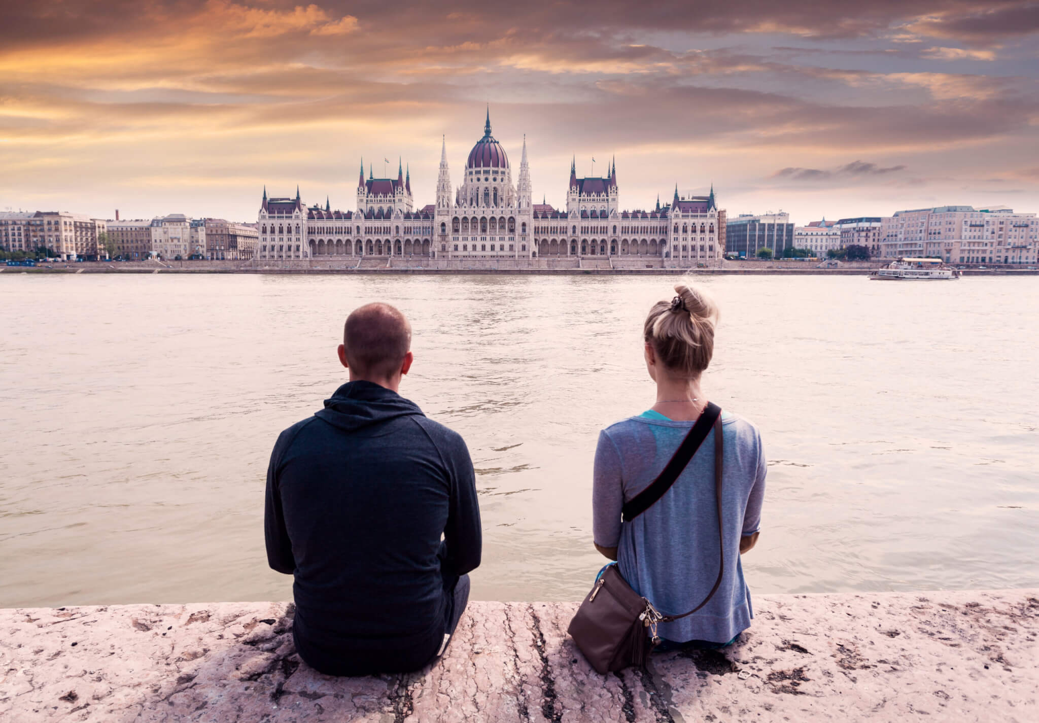 A couple in love sits on the embankment and enjoys the view of Parliament against the background of a sunset in Budapest, Hungary
