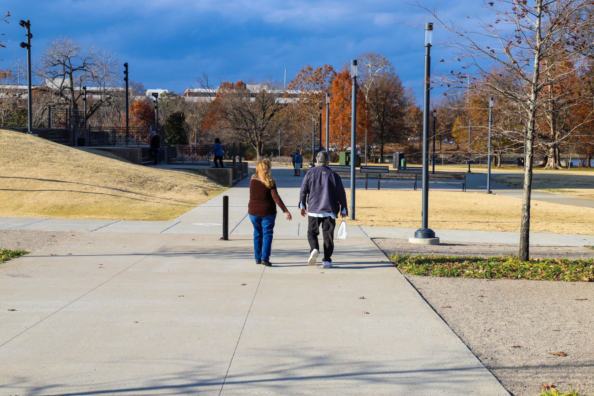 a couple walking down a footpath in a park with a stunning autumn landscape surrounded by autumn colored trees and grass with black light posts, blue sky and clouds at Centennial Park in Nashville