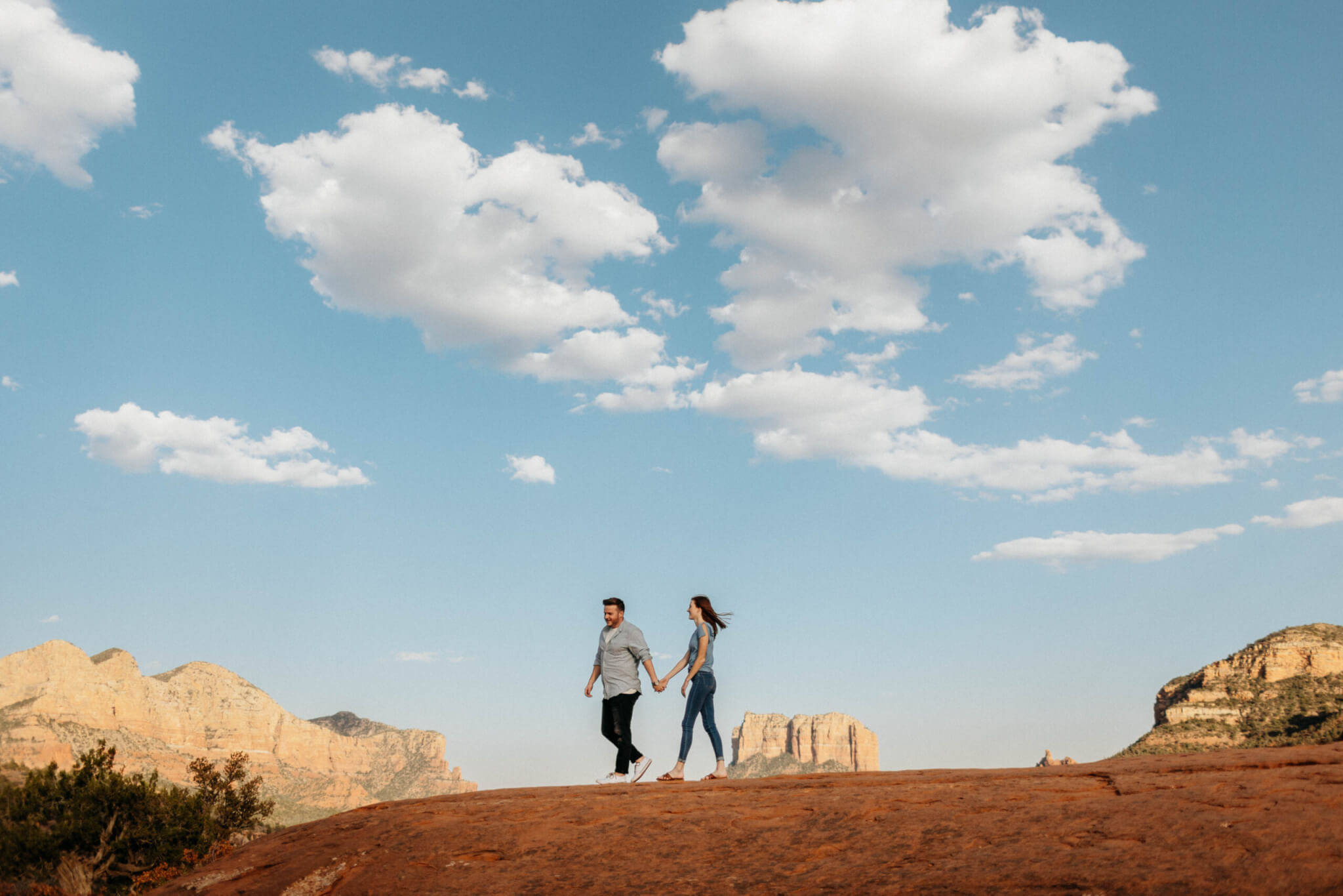 A man and woman holding hands standing while walking on brown rock formation under blue sky and white clouds in sedona arizona