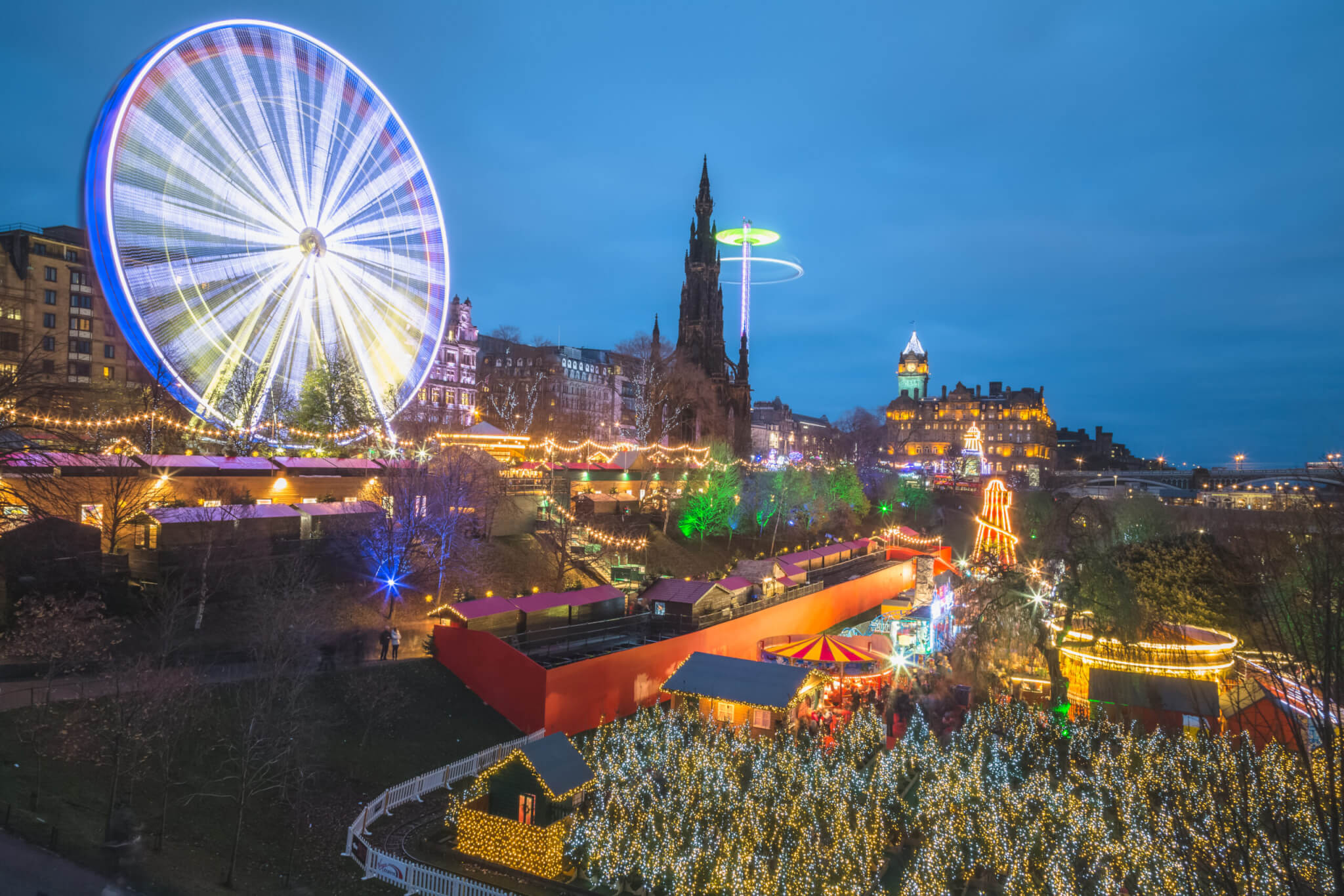 A vibrant and lively view of the Edinburgh Christmas Market at Princes Street Gardens in Scotland at night all lit up and illuminated with Christmas lights.