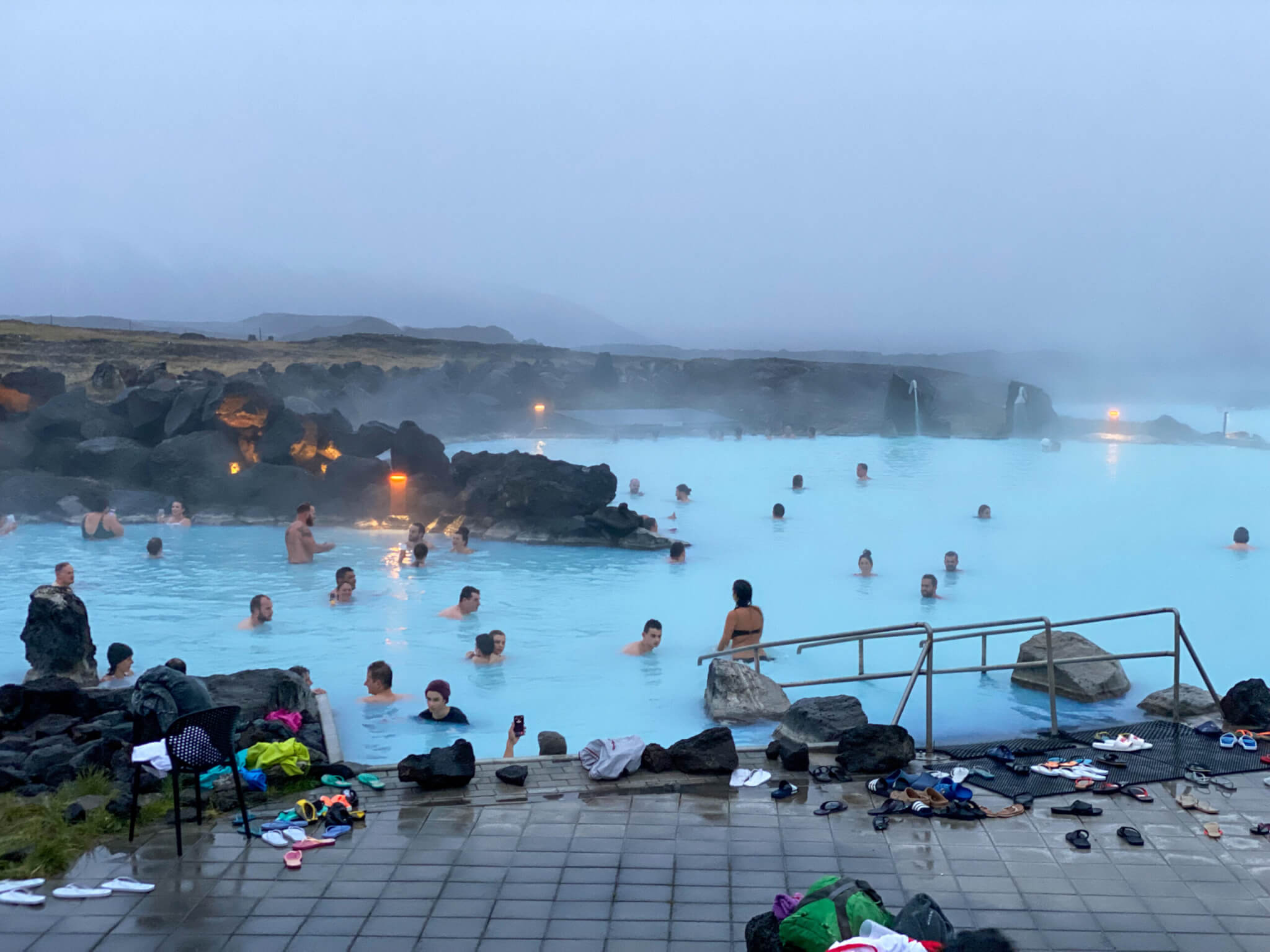 Myvatn Nature Baths in Iceland. Sky blue water with fog overhead, people in the water, gold lights on rocks.