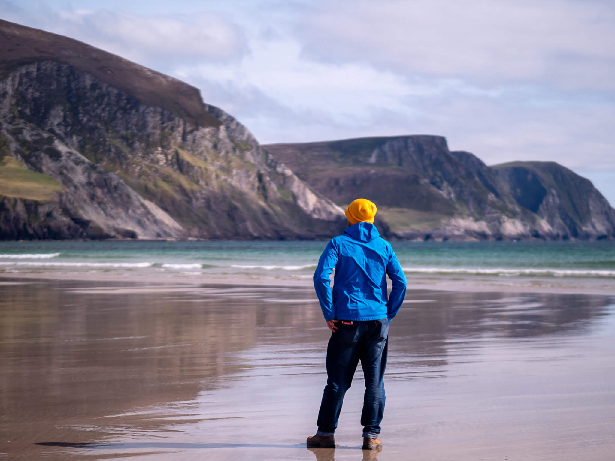 Slim man in blue jacket and yellow hat walking on a wet sand of a beach with beautiful nature scenery in the background. Keel beach, Ireland. Travel and tourism concept. Male on vacation by water.