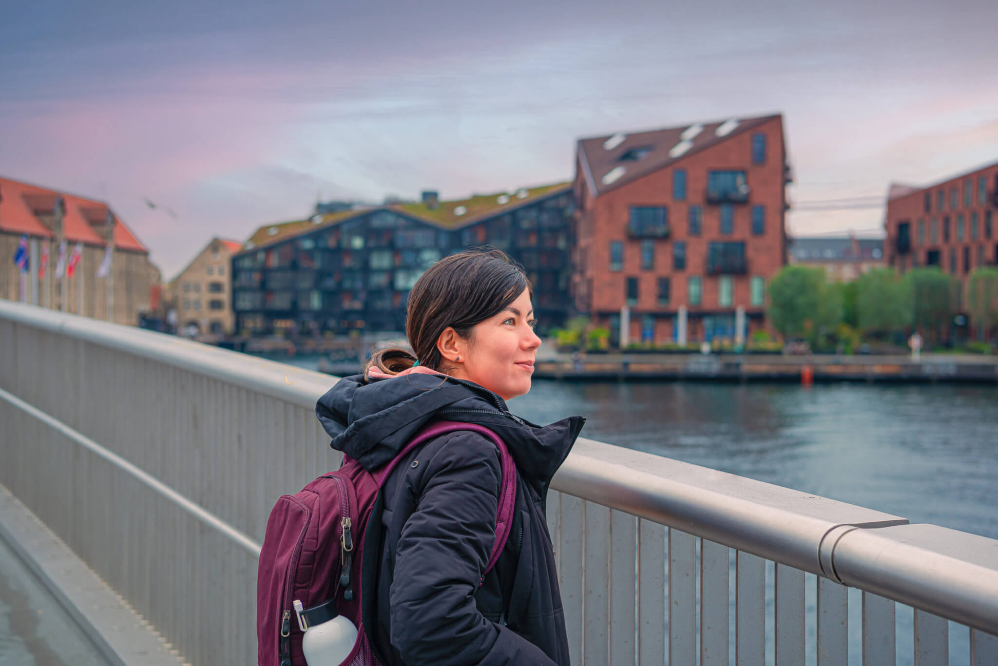 Young black-haired tourist woman  looks at the water of the Copenhagen canal, standing on the Inner Harbour pedestrian and bicyclist bridge Inderhavnsbroen against the neighbourhood Christianshavn