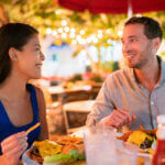 Couple eating hamburgers at outdoor restaurant terrace happy tourists on summer vacation. Florida travel people eating food at night during holidays in Miami. Asian Caucasian interracial young adults.