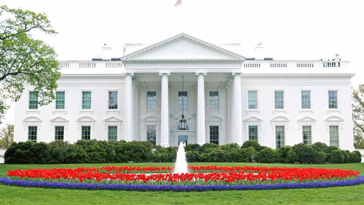 picture of the white house during the day time with red and blue flowers surrounding a fountain