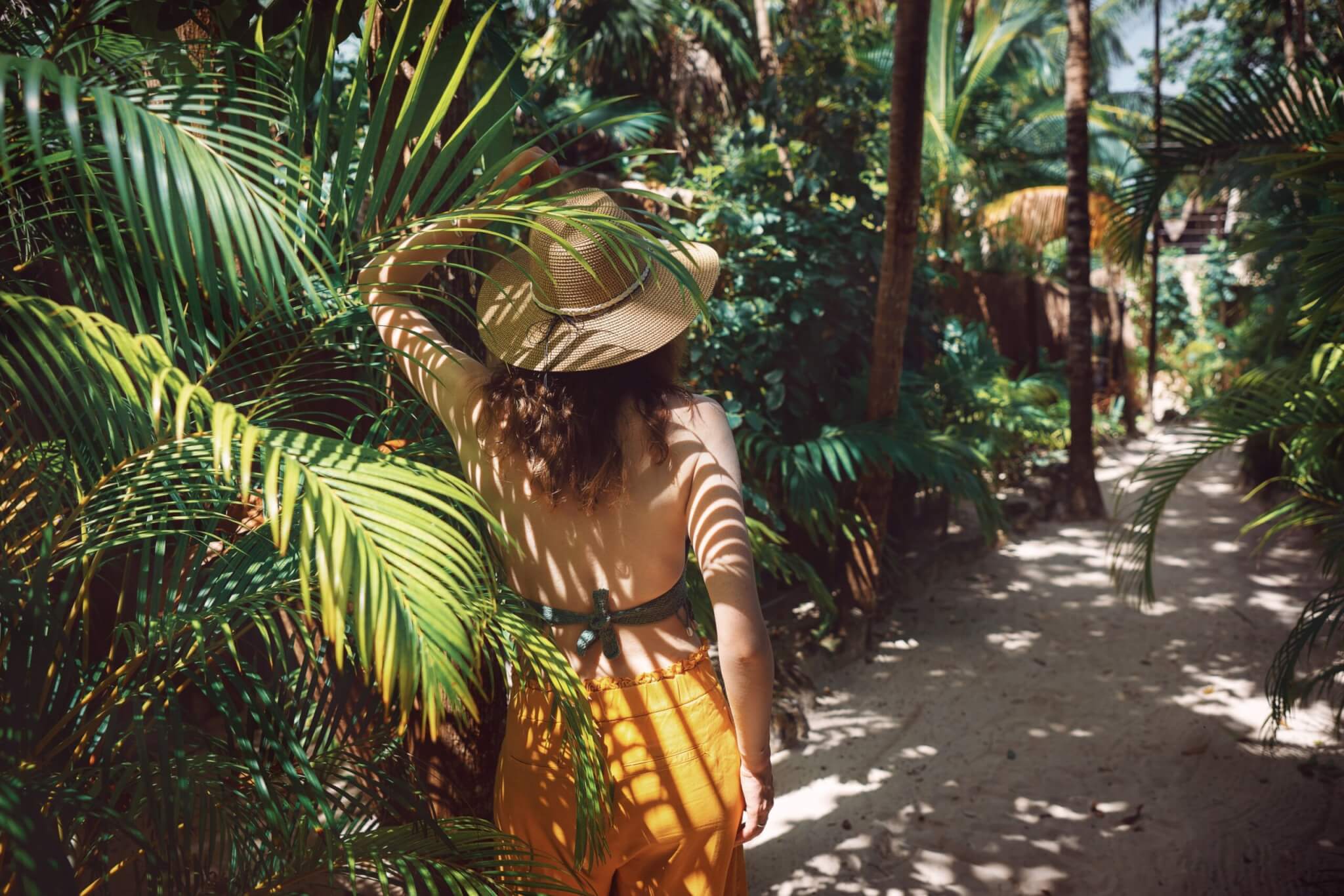 Young beautiful woman stands in the shade of palm trees, view from the back. A girl walks among tropical greenery on a bright sunny day in Mexico, Tulum.