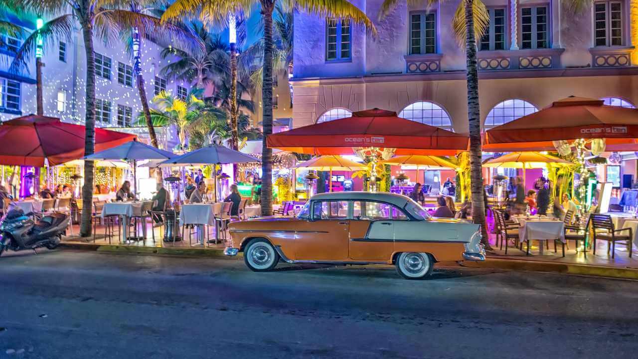 an old car is parked in front of a restaurant with umbrellas and palm trees