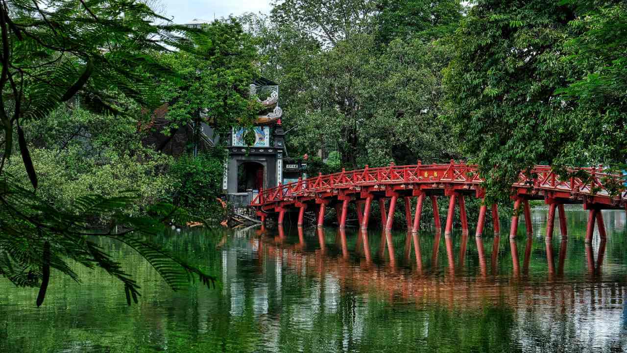 a red bridge over the water in the middle of a green park