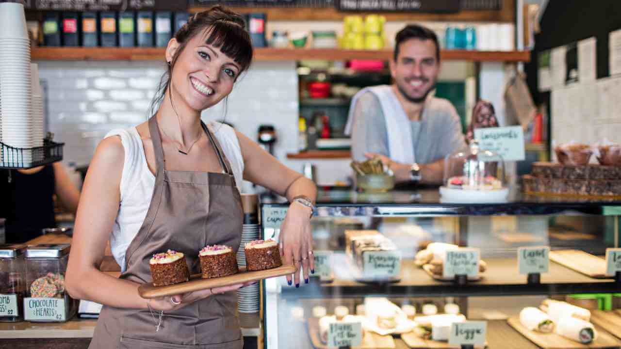 A person holding a tray of cupcakes in front of a bakery counter