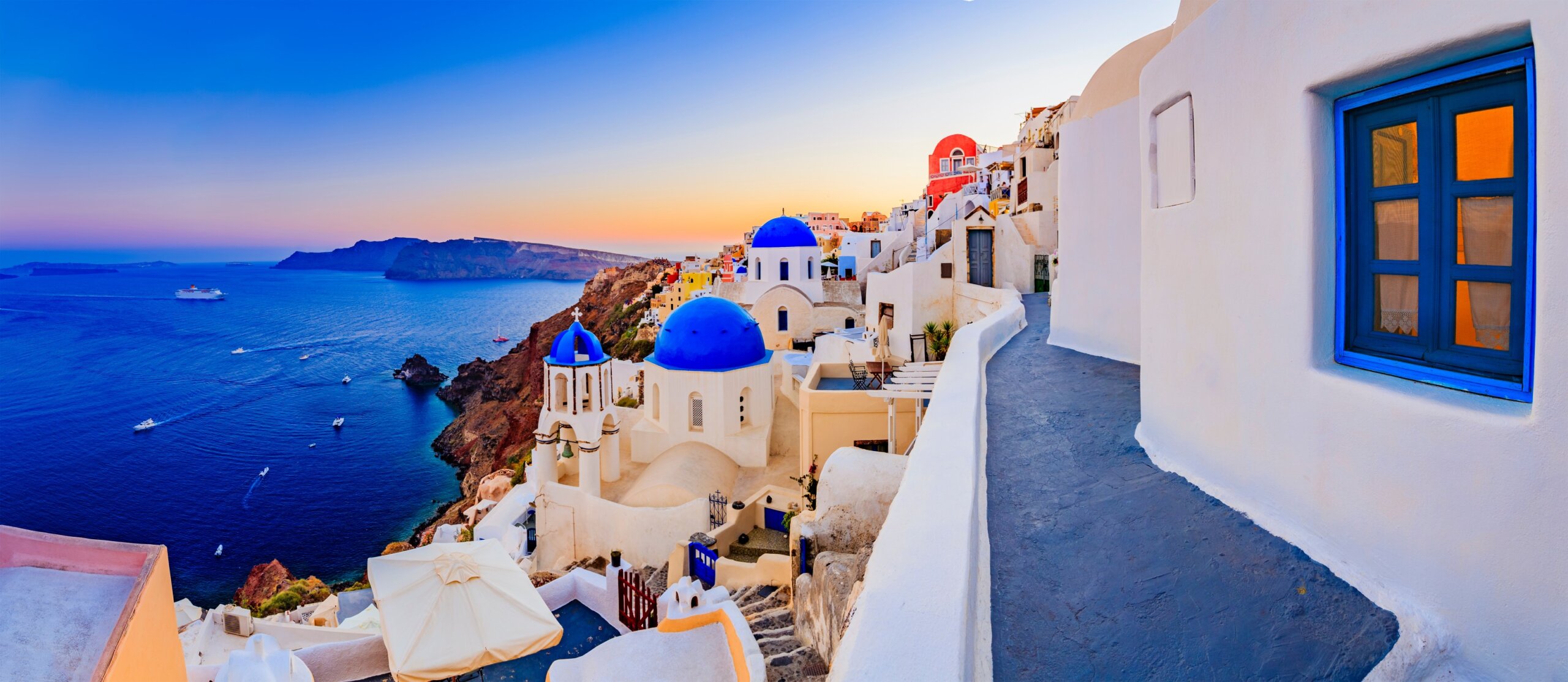 Panoramic sunset view of Oia, Santorini with whitewashed buildings, blue domes, and golden skies in September.