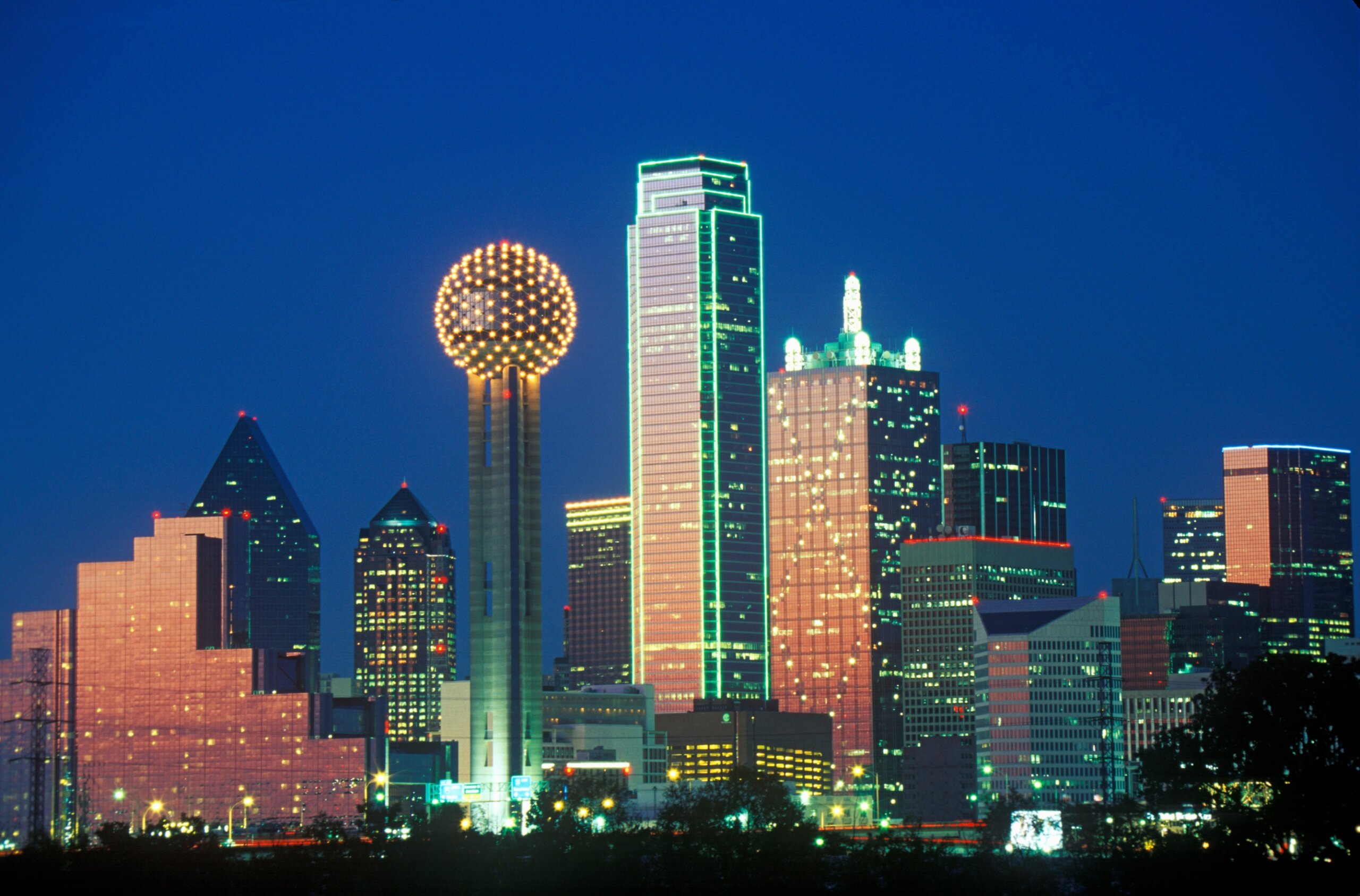 Nighttime view of Dallas, Texas skyline with brightly lit buildings and Reunion Tower glowing against the dark sky.
