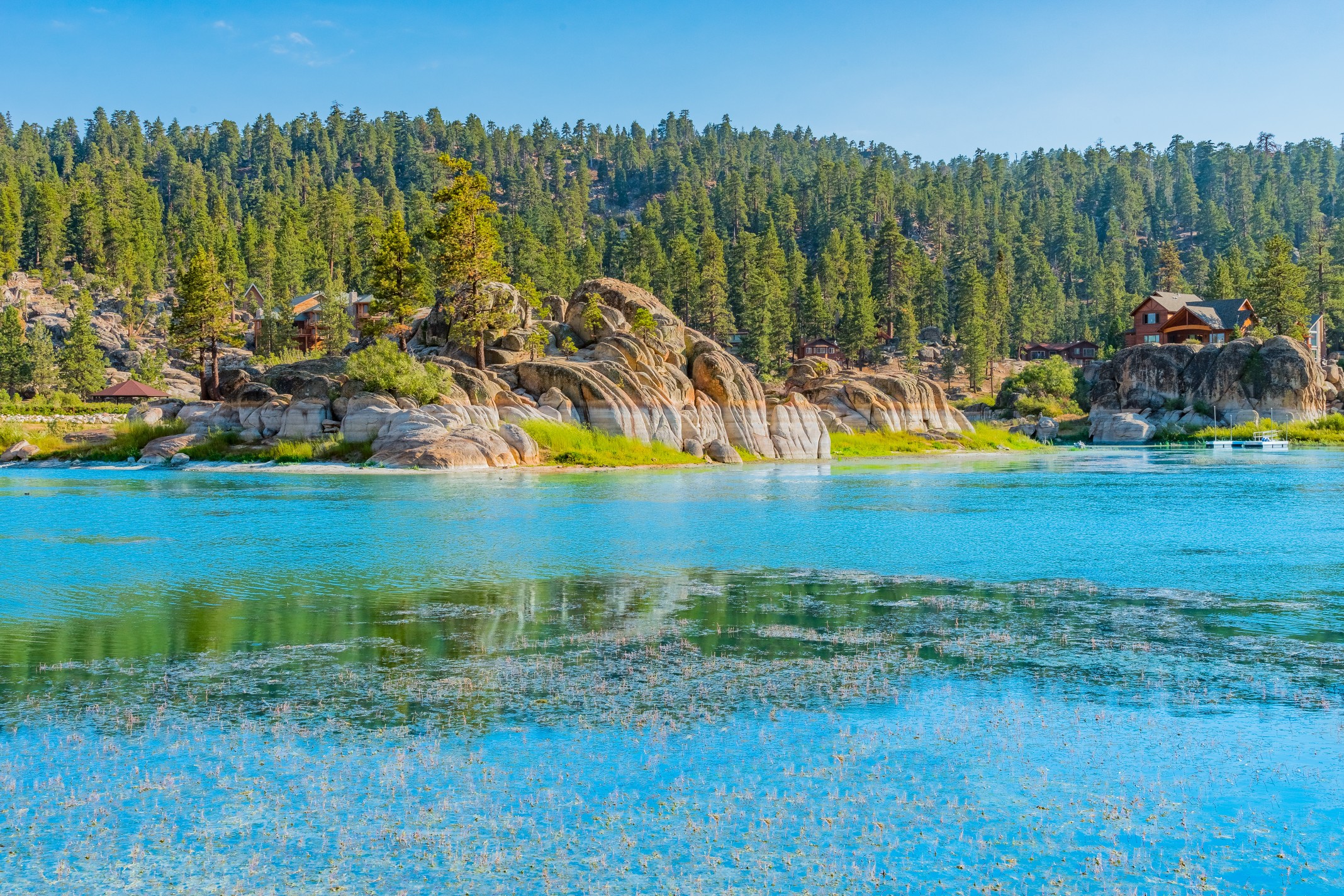 Colorful view of Boulder Bay at Big Bear Lake, with calm water, trees, and rocky shoreline under a clear sky.