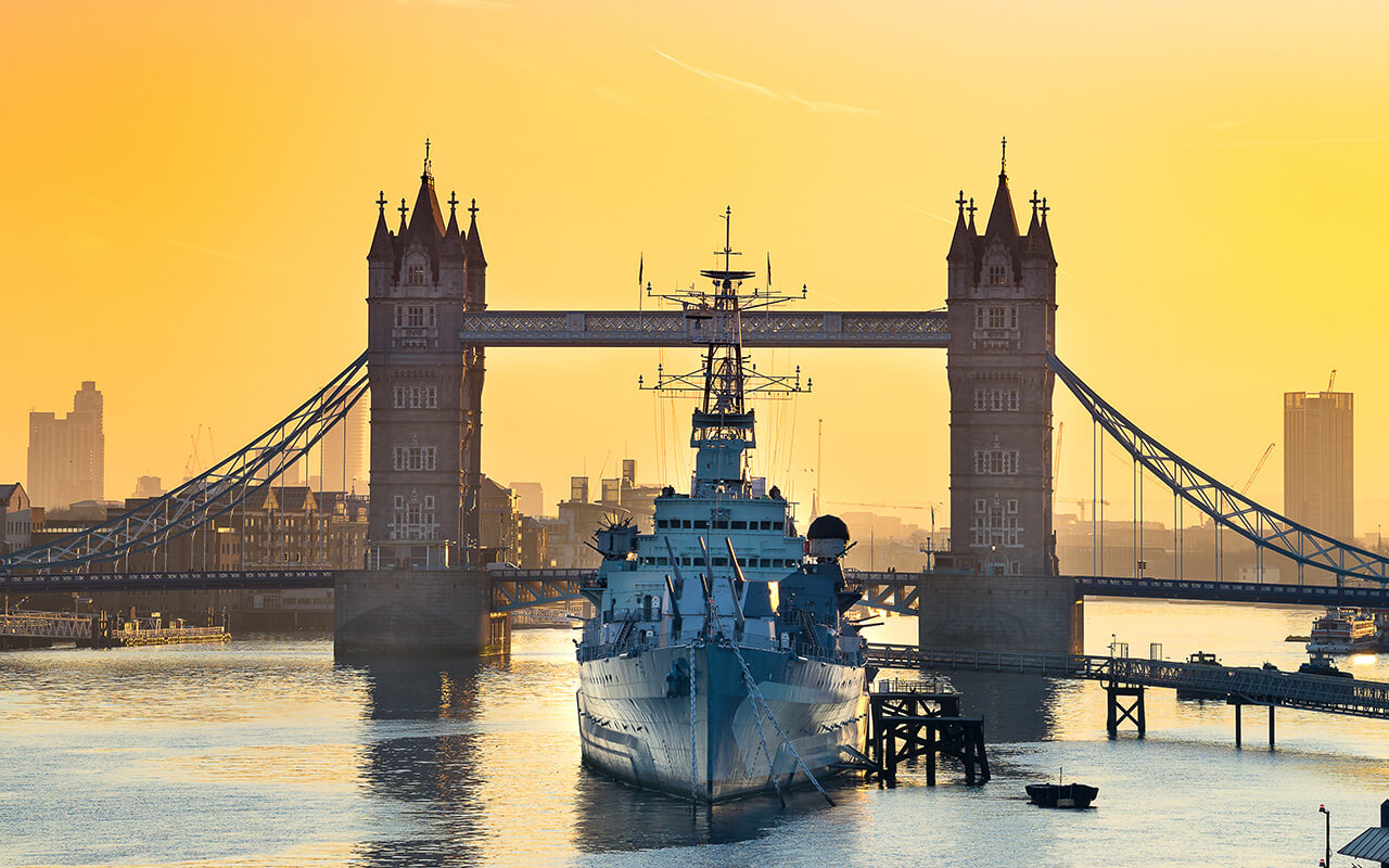 HMS Belfast moored in front of Tower Bridge on the River Thames at sunrise.