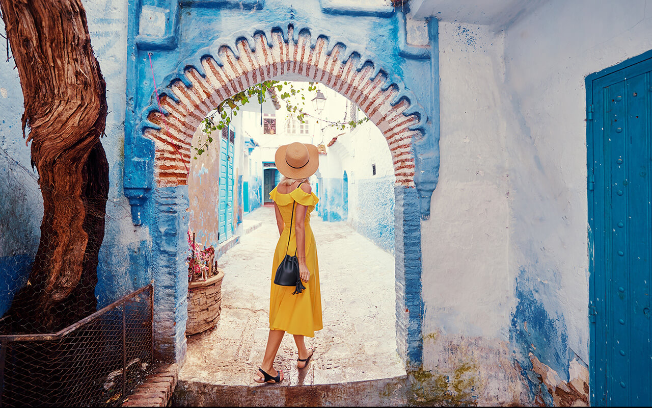 Colorful traveling by Morocco. Young woman in yellow dress walking in medina of blue city Chefchaouen.