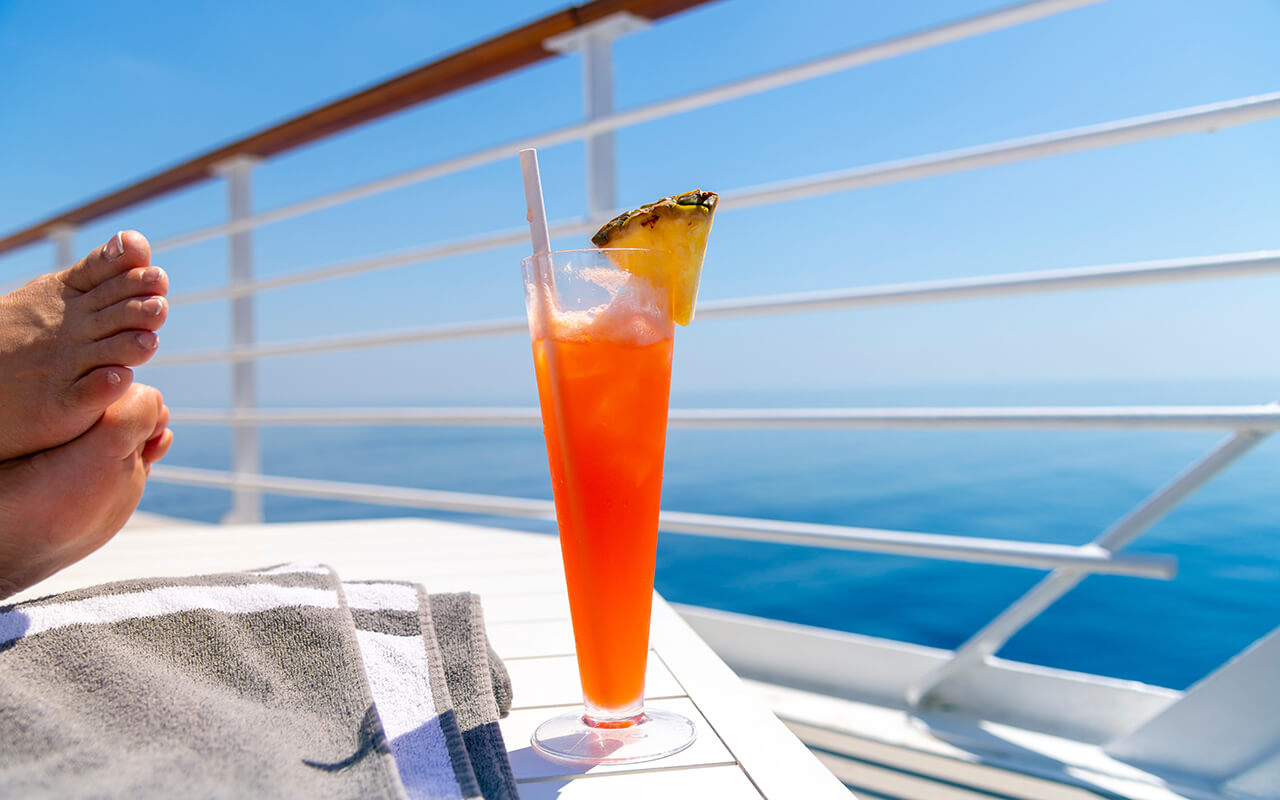 A woman rests her feet on a table on the upper deck of a cruise ship at sea with a colorful drink with pineapple on a hot summer day on the ocean.