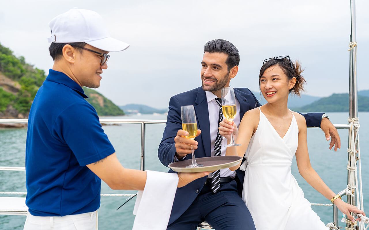 Asian man waiter serving champagne to passenger tourist while private catamaran boat yacht sailing in the ocean. 