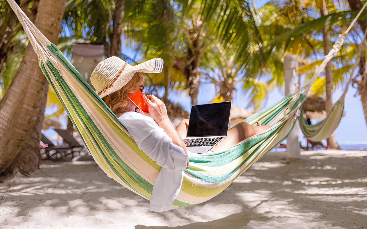 Woman lying in hammock on tropical beach and working on laptop and speaking on phone