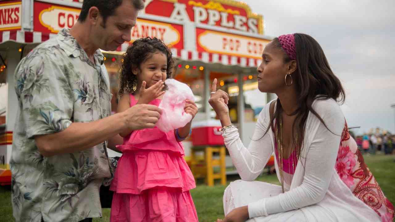 a couple with a child eating cotton candy in front of a carnival