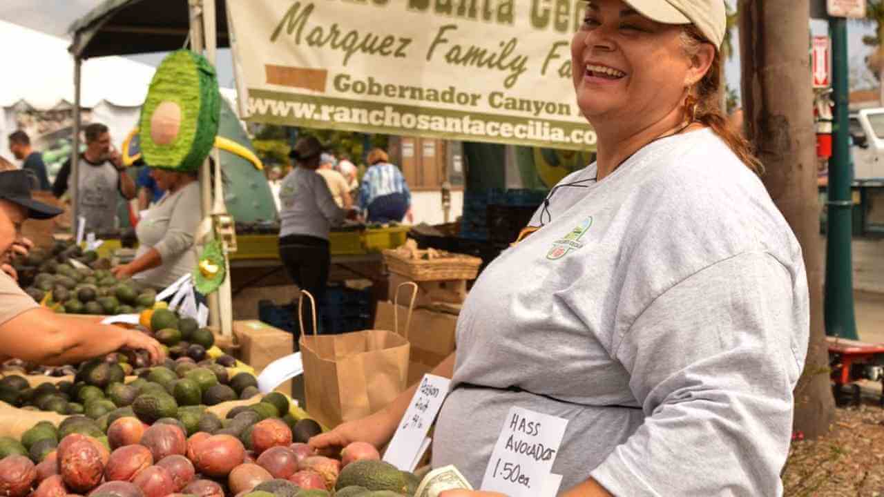 A person smiles as they hold avocados at a farmers market