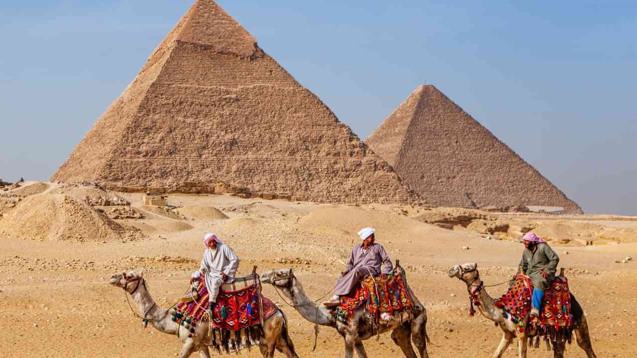 three people riding camels in front of the pyramids