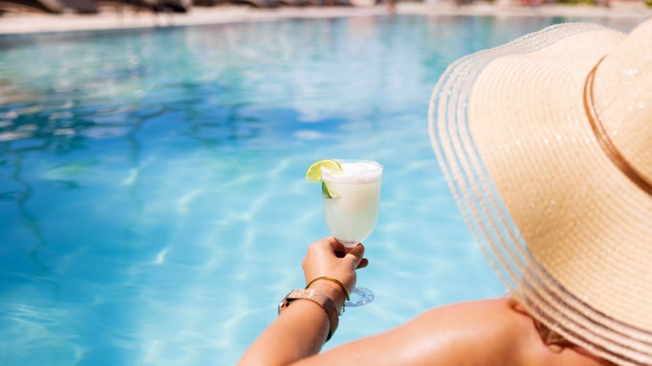 a person in a straw hat holding a drink in front of a swimming pool