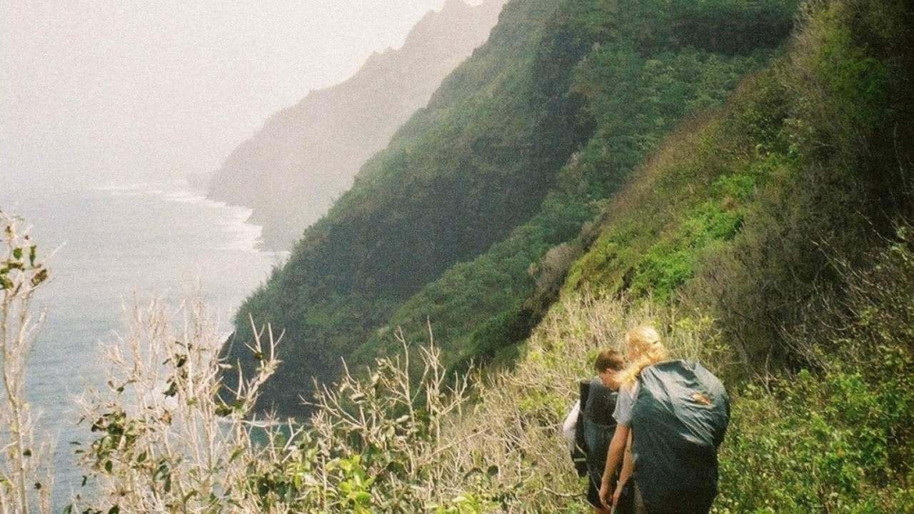 two people with backpacks walking along the side of a cliff overlooking the ocean