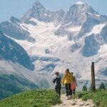 two people are walking up a mountain trail with snow capped mountains in the background