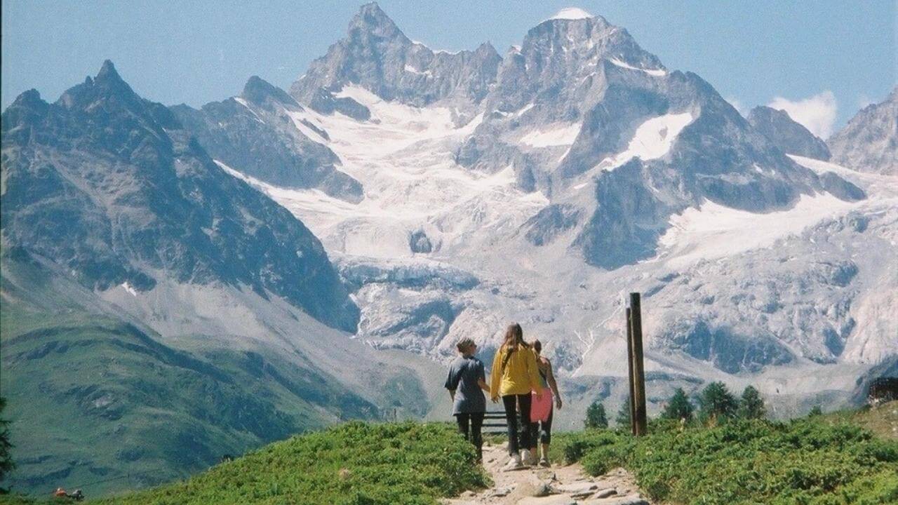 two people are walking up a mountain trail with snow capped mountains in the background