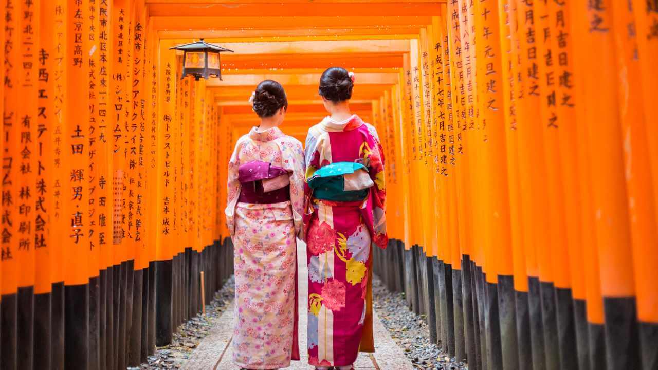 two individuals in traditional attire walking through a tunnel of orange tori gates