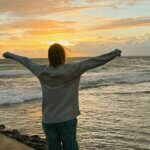 a person standing on the edge of a pier with their arms outstretched at sunset