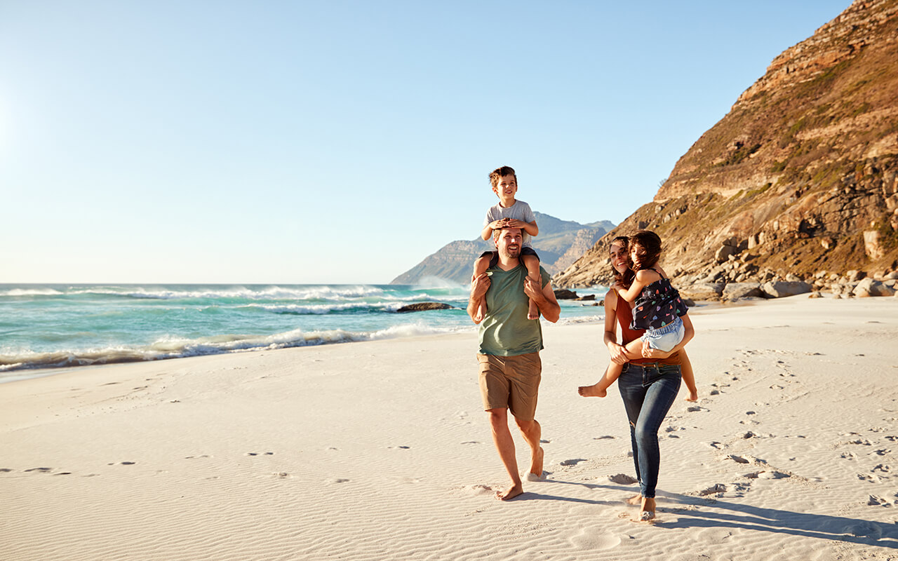 Mid adult white couple walking along on a beach on a family holiday carrying their kids, full length
