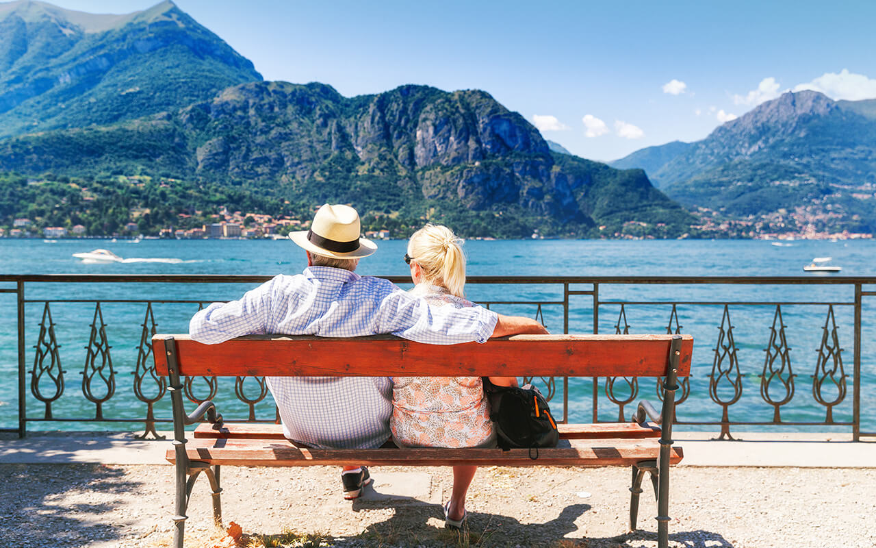 Senior couple sitting on a bench overlooking mountains and lake while traveling – travel tips for seniors
