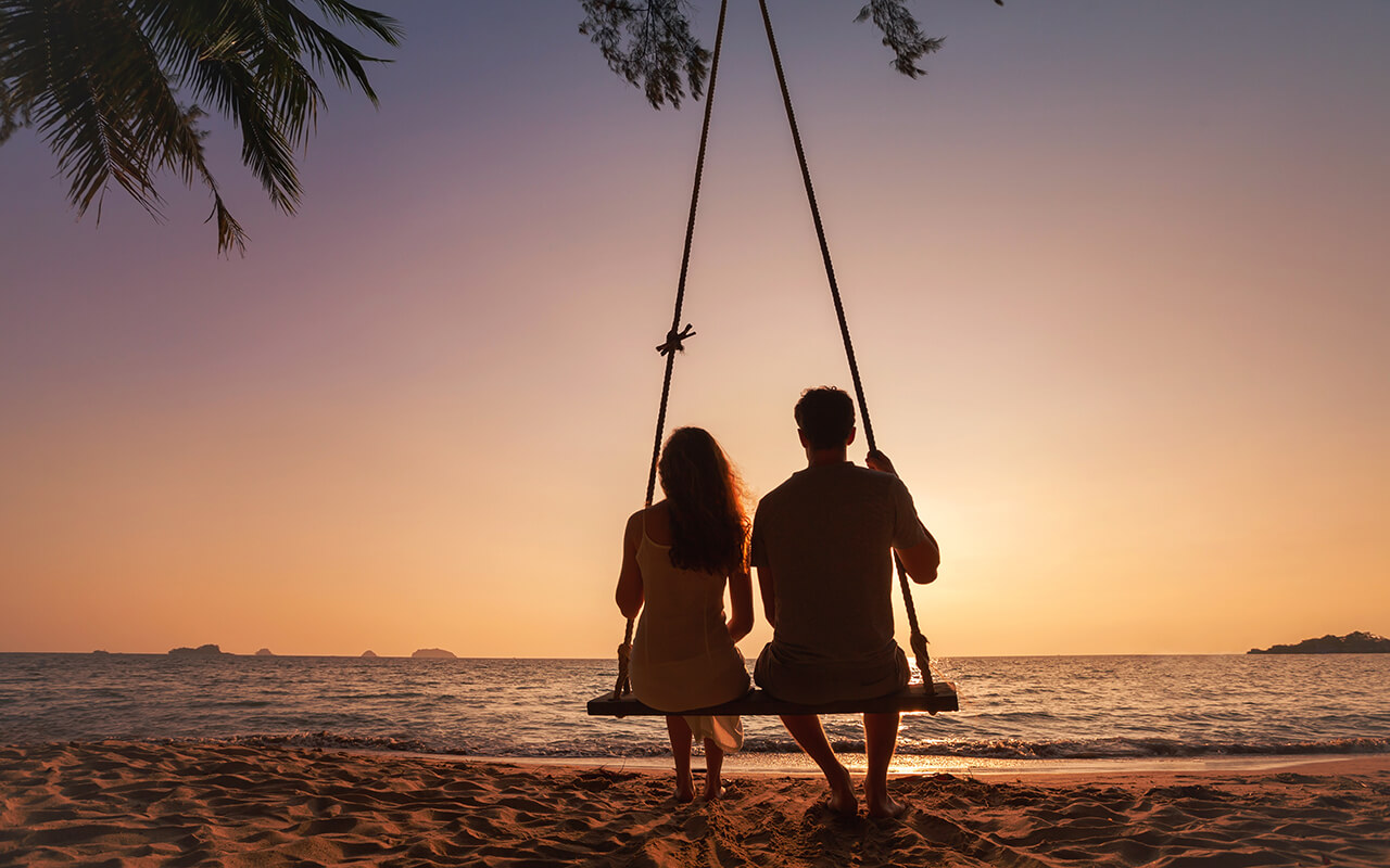 Couple sitting on a beach swing at sunset looking at the ocean on a romantic escape