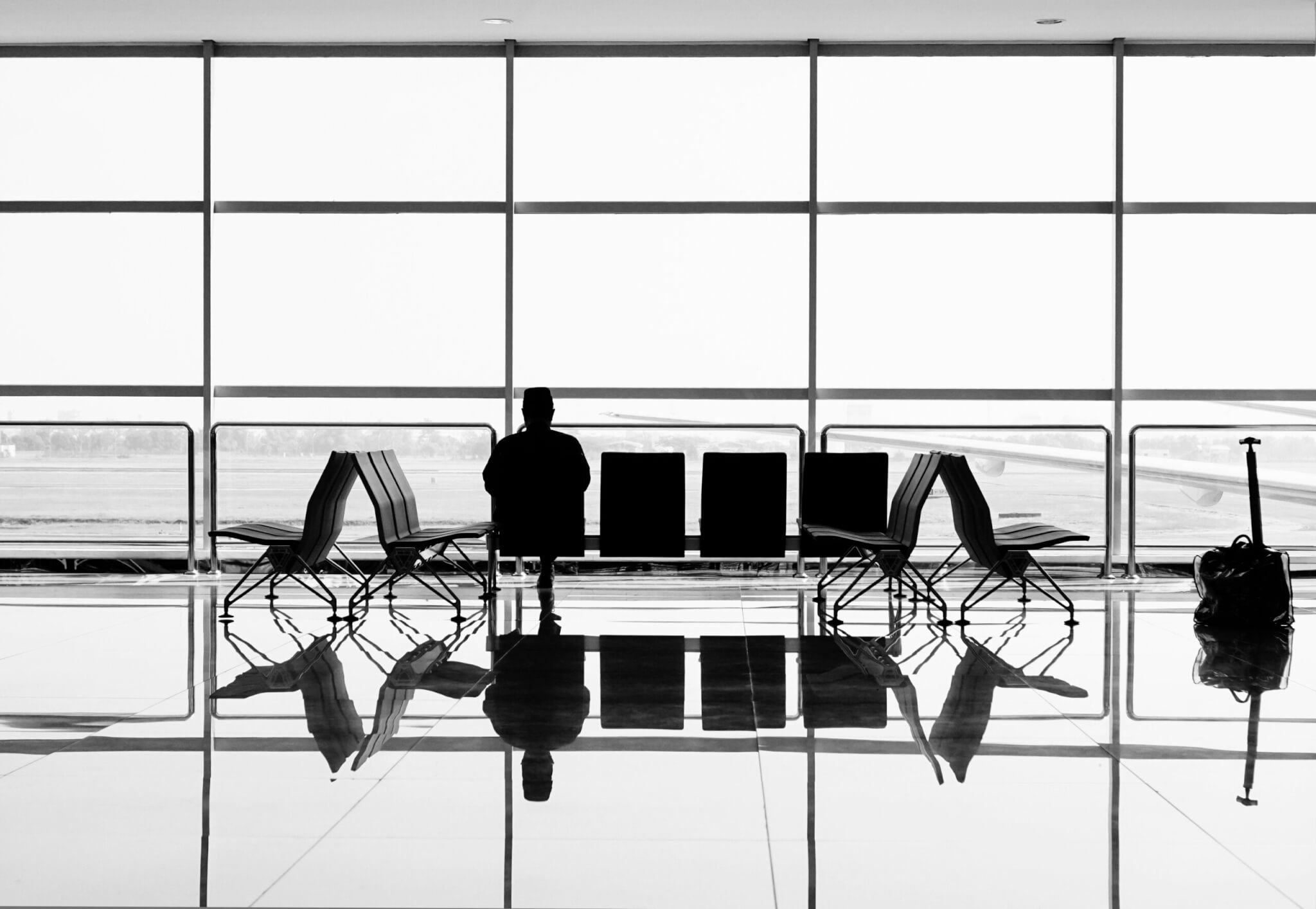 Traveler sitting alone in an airport terminal with large windows and empty chairs.