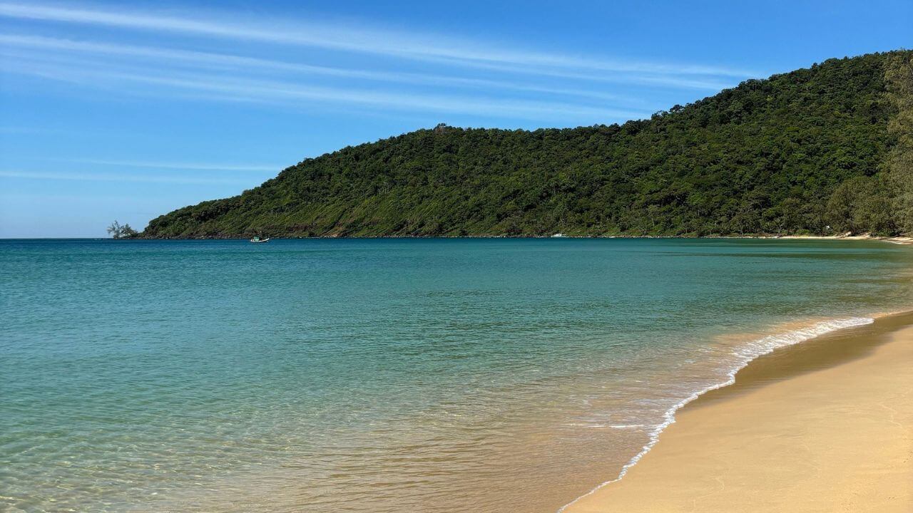 a sandy beach with clear water and green hills in the background